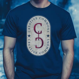 以下が含まれることがあります： ネイビーブルーのTシャツ。中央に「Creative Commons」の文字と赤い「CC」の文字、2つの星が描かれた、使い古された楕円形のデザイン。柔らかい素材で作られたTシャツです。