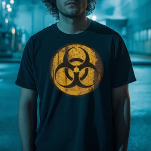 以下が含まれることがあります： ネイビーブルーのTシャツ。黄と黒のバイオハザードマークがプリントされています。円形のグラフィックは使い古されたヴィンテージ風です。Tシャツはクルーネックです。
