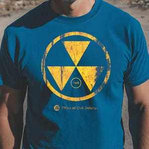 May include: A blue t-shirt featuring a yellow radiation symbol design. The design includes three triangles within a circle, with the number "1500" inside a smaller circle. The text "Office of Civil Defense" is below the symbol.