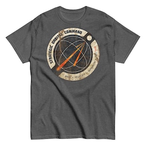 May include: Dark gray t-shirt featuring a distressed circular graphic with the text "STRATEGIC ORBITAL COMMAND" and "MASS VELOCITY AUTHORITY". The graphic includes an orange arrow design over a globe.