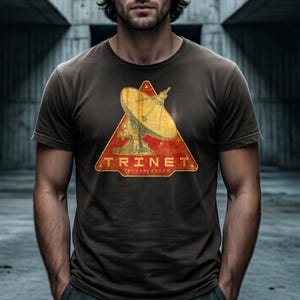 Pode incluir: T-shirt castanho escuro com um gráfico desgastado de uma antena parabólica dentro de um triângulo vermelho. O texto "TRINET TECHNOLOGIES" é impresso abaixo em estilo vintage.