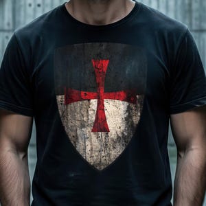 Könnte beinhalten: Schwarzes T-Shirt mit einem verwitterten Schilddesign und einem roten Kreuzemblem. Das Schild hat ein abgenutztes Aussehen mit Schwarz-, Weiß- und Brauntönen. Das Shirt ist kurzärmlig und aus dunklem Stoff gefertigt.