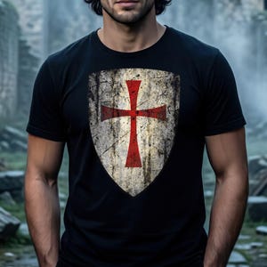 May include: A black short-sleeved t-shirt featuring a large, centered graphic of a weathered, distressed shield. The shield design displays a prominent red cross against a textured, off-white and grey background, mimicking an aged metal surface.