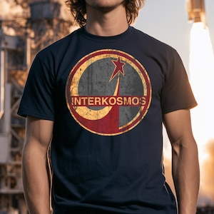 May include: A dark navy blue short-sleeve t-shirt featuring a circular graphic on the chest. The design includes a distressed red and grey emblem with a star and the text INTERKOSMOS printed in a bold, sans-serif font across the center.