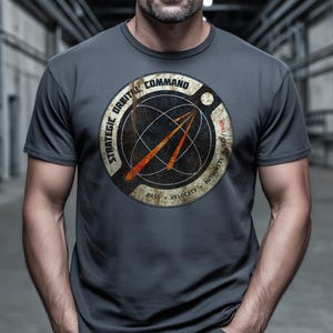 Op de afbeelding: Grijs t-shirt met een verweerde cirkelvormige afbeelding. De afbeelding bevat de tekst "STRATEGIC ORBITAL COMMAND" en "MASS VELOCITY AUTHORITY", samen met een wereldbolontwerp en oranje pijlen.