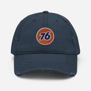Könnte beinhalten: Eine abgenutzte Baseballkappe in Marineblau mit einem weißen und orangefarbenen gestickten Kreis, der die Zahl "76" in Marineblau enthält.