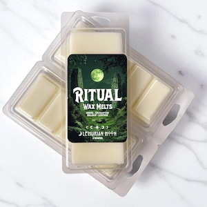 May include: Two clear plastic clamshell containers filled with off-white wax melts. The center label features a green forest scene with a full moon, the text RITUAL WAX MELTS, GRASS EUCALYPTUS BALSAM LEATHER, and LEMURIAN MOON with 2 OUNCES printed below.