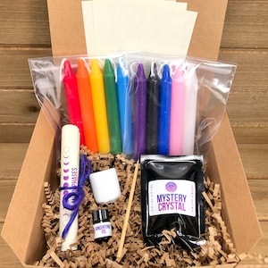 May include: A cardboard box filled with a variety of items for a magical ritual. The box contains a set of rainbow colored candles, a small bottle of anointing oil, a white ceramic dish, a small bag of black crystals labeled "Mystery Crystal", a rolled up piece of paper with the words "Moon Phases" written on it, and a wooden skewer.