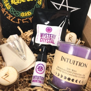 May include: A gift box filled with items for a spiritual practice. The box contains a purple candle with the word "INTUITION" on the label, a small bottle of oil labeled "INTUITION ANOINTING OIL", a small bag labeled "MYSTERY CRYSTAL", a clear crystal point, and two wooden runes.