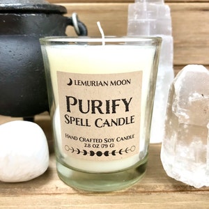 May include: A white soy candle in a glass jar with a brown label that reads "Lemurian Moon Purify Spell Candle Hand Crafted Soy Candle 2.8 oz (79 g)".