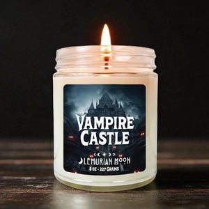 Puede incluir: Una vela encendida en un tarro de cristal con una etiqueta que dice "Vampire Castle" sobre una ilustración de un castillo oscuro. La etiqueta también dice "Lemurian Moon" y la vela pesa 227 gramos.