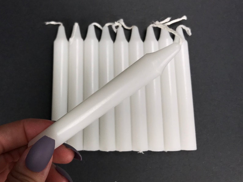 May include: A bundle of ten white taper candles, each approximately 7 inches long. The candles are arranged in a fan shape, with the tips of the candles pointing towards the right side of the image.