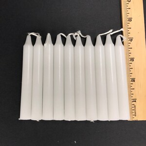 May include: Ten white taper candles, each approximately 3 inches long, are lined up next to a wooden ruler.