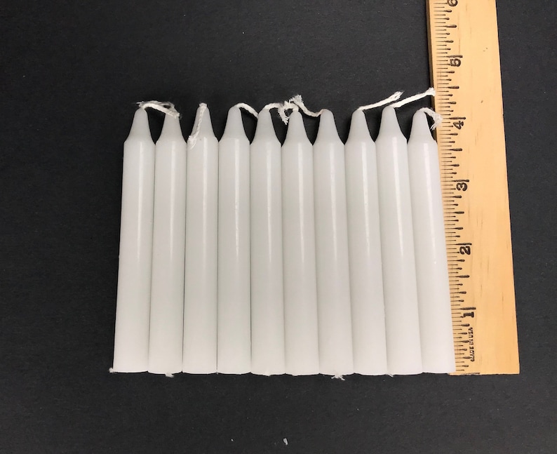May include: Ten white taper candles, each approximately 3 inches long, are lined up next to a wooden ruler.
