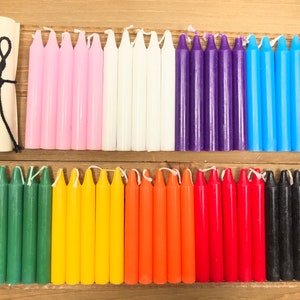 May include: A set of 70 small, thin candles in various colors, including pink, white, purple, blue, green, yellow, orange, red, and black. The candles are arranged in rows on a wooden surface.