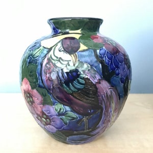 Könnte beinhalten: Eine runde Keramikvase mit einem farbenfrohen Design, das einen Vogel mit langem Schwanz, Blumen und Trauben zeigt. Die Vase ist in Blautönen, Lila, Grün und Gelb bemalt.
