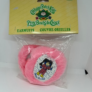 May include: Pink Cabbage Patch Kids earmuffs with a white patch featuring a Cabbage Patch Kid wearing a blue and red checked outfit and blue earmuffs. The text "Cabbage Patch Kids" is printed on the patch.