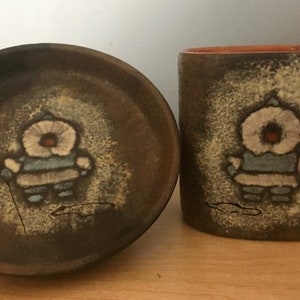 Peut inclure: Un ensemble de deux plats en céramique brune avec un motif fantaisiste d'une personne vêtue d'une tenue bleue et blanche. Les plats ont un aspect rustique et peint à la main.