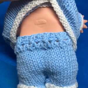 Pu&ograve; includere: Un completo di maglia blu per una bambola per bambini. Il completo comprende un maglione a maniche lunghe e pantaloncini con bordini bianchi.