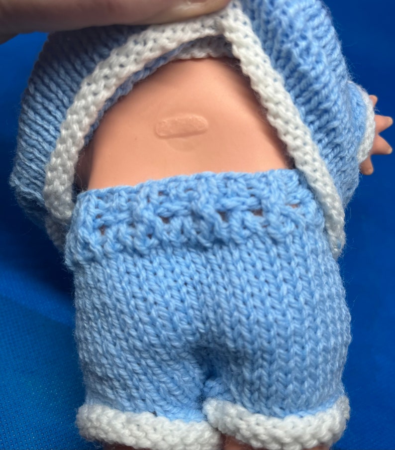 Pu&ograve; includere: Un completo di maglia blu per una bambola per bambini. Il completo comprende un maglione a maniche lunghe e pantaloncini con bordini bianchi.
