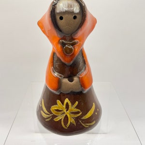 Op de afbeelding: Een keramiek beeldje van een vrouw die een rode mantel met capuchon en een bruine jurk met een gouden bloemenmotief draagt. Het beeldje heeft een bruin gezicht met zwarte ogen en wit haar.