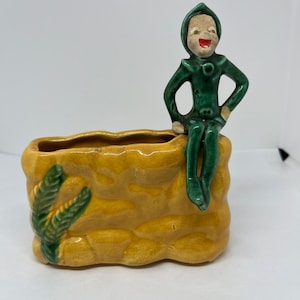 Könnte beinhalten: Ein Keramik-Pflanztopf in Form einer Steinmauer mit einer grünen Elfenfigur, die oben sitzt. Der Topf hat eine kleine Öffnung zum Pflanzen und ist mit grünen Blättern verziert.