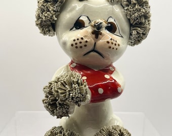 May include: A white ceramic poodle figurine with brown eyes and a red and white polka dot scarf. The poodle has gray pom-poms for fur.