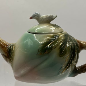 May include: A small, green and brown teapot with a bird-shaped lid. The teapot is decorated with gold leaf and has a swirling design.