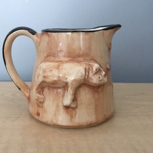May include: A small, brown ceramic creamer with a black rim and handle. The creamer features a raised design of a pig on the front.
