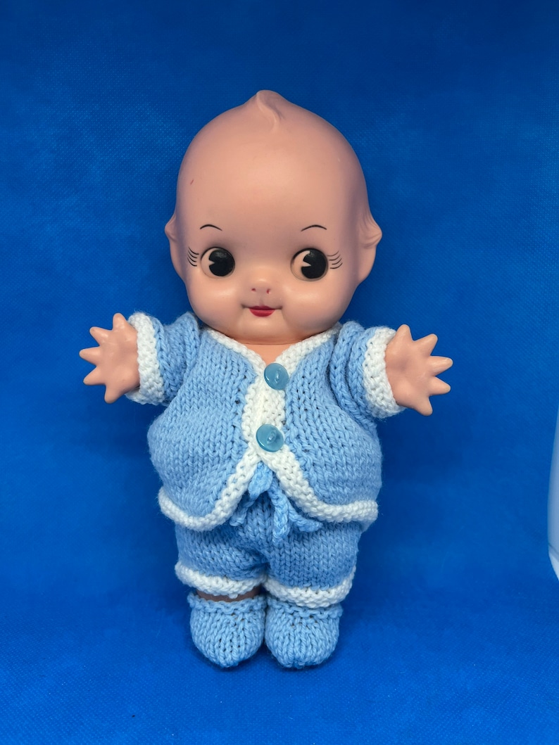 Pu&ograve; includere: Una bambola di porcellana vintage che indossa un completo di maglia blu con rifiniture bianche. La bambola ha occhi grandi e un sorriso dipinto.