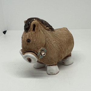 Pode incluir: Uma figura de burro de cerâmica marrom com cascos brancos e uma flor azul no pescoço. O burro tem uma juba marrom e uma superfície texturizada.