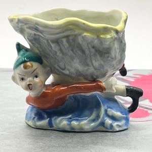 Peut inclure: Une figurine en céramique d'un petit lutin tenant une grande coquille de mer grise et jaune. Le lutin porte un chapeau vert, une chemise orange, un pantalon bleu et des chaussures noires. La coquille a un intérieur blanc et est en forme de palourde.
