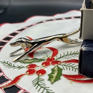 Op de afbeelding: Een zilverkleurig, gestileerd hondenbeeldje in een rennende pose. Het beeldje staat op een wit kantje met rode en groene borduursels.