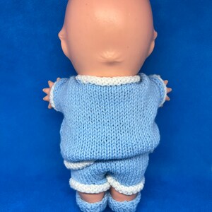 Pu&ograve; includere: Un completo di maglia blu per una bambola, con bordino bianco intorno al collo, alle maniche e ai pantaloncini. Il completo &egrave; realizzato con un filato morbido e testurizzato.