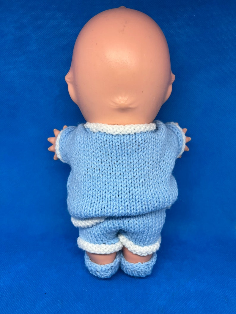 Pu&ograve; includere: Un completo di maglia blu per una bambola, con bordino bianco intorno al collo, alle maniche e ai pantaloncini. Il completo &egrave; realizzato con un filato morbido e testurizzato.