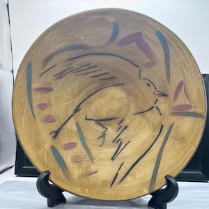 May include: A decorative plate featuring a bird design. The plate has a tan background with a black bird illustration. Blue, purple, and pink accents are painted around the bird. The plate is displayed on a black stand.