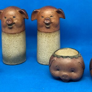 Może przedstawiać: Zestaw pięciu ceramicznych figurek świń. Dwa wysokie solniczki i pieprzniczki w kształcie świń z brązową i beżową glazurą. Trzy mniejsze figurki świń z brązową i beżową glazurą.