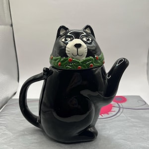 Könnte beinhalten: Schwarze Keramik-Teekanne in Form einer Katze, die einen grünen Stechpalmenkranz mit roten Beeren um den Hals trägt.