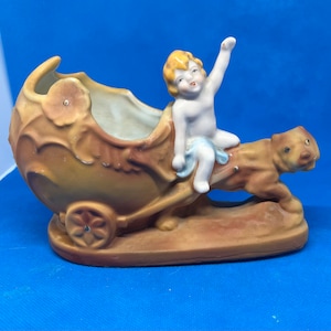 May include: A porcelain figurine of a cherub riding a chariot pulled by a dog. The chariot is a planter with a brown and cream glaze. The cherub is wearing a blue dress and has blonde hair.