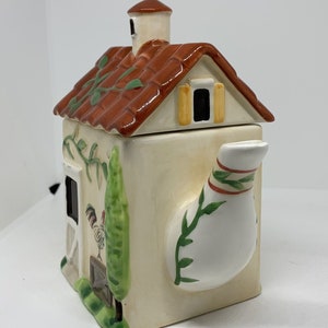 Peut inclure: Une théière en céramique blanche en forme de maison avec un toit brun, des vignes vertes et une illustration de coq. La théière a un bec en forme de cheminée et une poignée rayée rouge et verte.