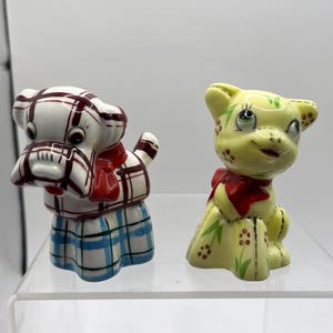 May include: Two ceramic animal ornaments. One is a white dog with a red and white plaid pattern, a red scarf, and a blue and white plaid skirt. The other is a yellow cat with a red bow, green leaves, and red berry accents.