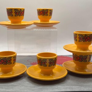 May include: Set of six vintage yellow ceramic egg cups with matching saucers. Each cup features a decorative band of red berries and green leaves. The egg cups are a bright, cheerful addition to any breakfast table.