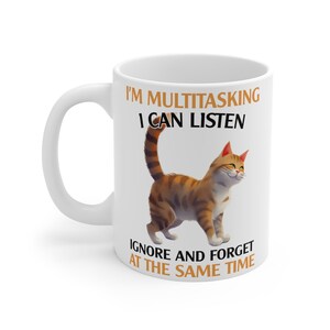 Pode incluir: Caneca de cerâmica branca com uma ilustração de um gato tigrado marrom. A caneca diz "I'm multitasking I can listen ignore and forget at the same time."