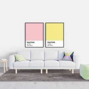 May include: A modern living room scene featuring a white sofa, a small wooden side table, and a green ottoman. Two framed Pantone color charts hang on the wall above the sofa, one pink and one yellow. A gray rug sits beneath the sofa.