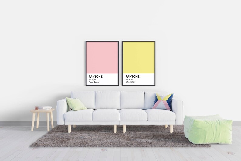 May include: A modern living room scene featuring a white sofa, a small wooden side table, and a green ottoman. Two framed Pantone color charts hang on the wall above the sofa, one pink and one yellow. A gray rug sits beneath the sofa.