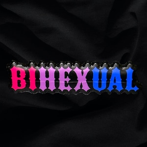 May include: A black fabric background with a colorful patch that reads "Bisexual" in a bold, stylized font. The letters are outlined in black and filled with pink, purple, and blue.