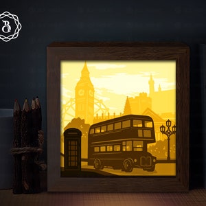 Może przedstawiać: Żółta wycinanka papierowa przedstawiająca londyński autobus piętrowy, Big Ben i inne ikoniczne zabytki. Praca jest oprawiona w brązową drewnianą ramę.