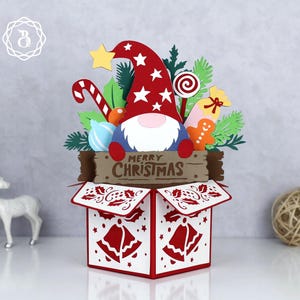 May include: A festive Christmas-themed papercraft box featuring a gnome with a red hat and white stars. The box is decorated with red bells and holly. The words "MERRY CHRISTMAS" are displayed on a wooden sign.
