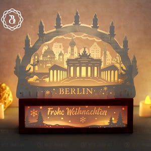 Puede incluir: Un adorno decorativo iluminado de papel recortado de los monumentos de Berlín, incluyendo la Puerta de Brandeburgo y la Torre de Televisión. El diseño es blanco y se asienta sobre una base roja con las palabras "BERLIN" y "Frohe Weihnachten" iluminadas.