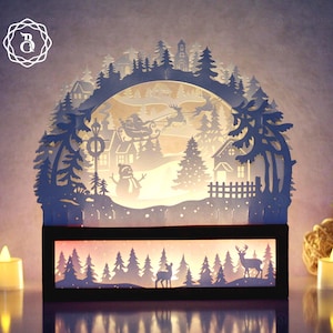 Könnte beinhalten: Eine mehrschichtige Weihnachtsszene aus Papier mit einem Winterdorf, Bäumen und Rentieren. Die Szene wird von innen beleuchtet und erzeugt einen warmen Schein. Die Basis ist ein schwarzes Rechteck mit einer Waldsilhouette.